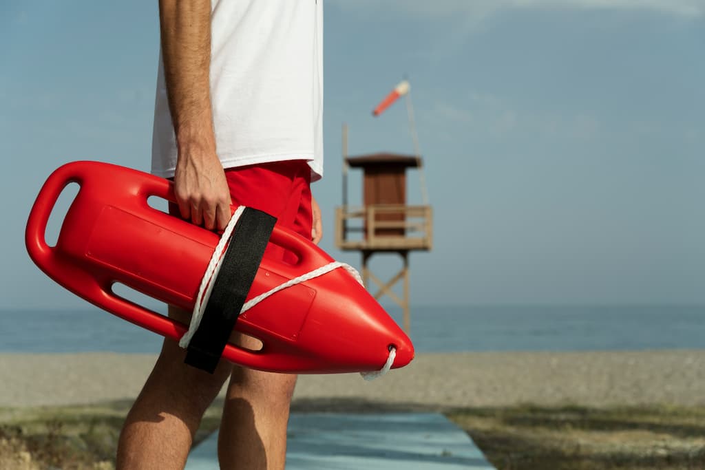 FotografĆa de un socorrista en la playa, utilizada para representar el proyecto LAIF de CIMNE dentro de la Unidad de Innovación DIGIT. El socorrista lleva unos pantalones cortos rojos y una camiseta blanca, sujetando una boya de rescate roja, con una torre de socorrista y una bandera roja visibles al fondo. La escena enfatiza la seguridad costera, la preparación para emergencias y la monitorización centrada en las personasātemas clave en la investigación de LAIF sobre sistemas inteligentes para el apoyo de socorristas y la gestión de playas.
