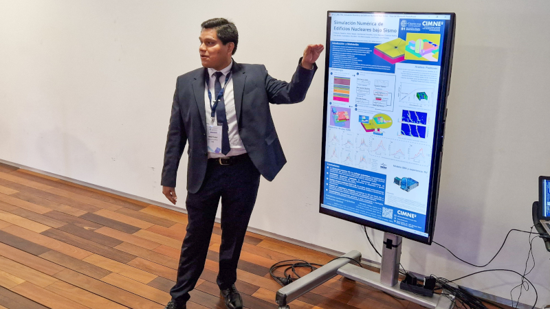 Cristhian Albert Padila presenting a scientific poster displayed on a vertical digital screen. The poster, from CIMNE, is titled “Simulación Numérica de Edificios Nucleares bajo Sismo” and includes colorful diagrams, graphs, and simulation results related to nuclear building performance under seismic conditions. The presentation takes place indoors on a wooden floor.