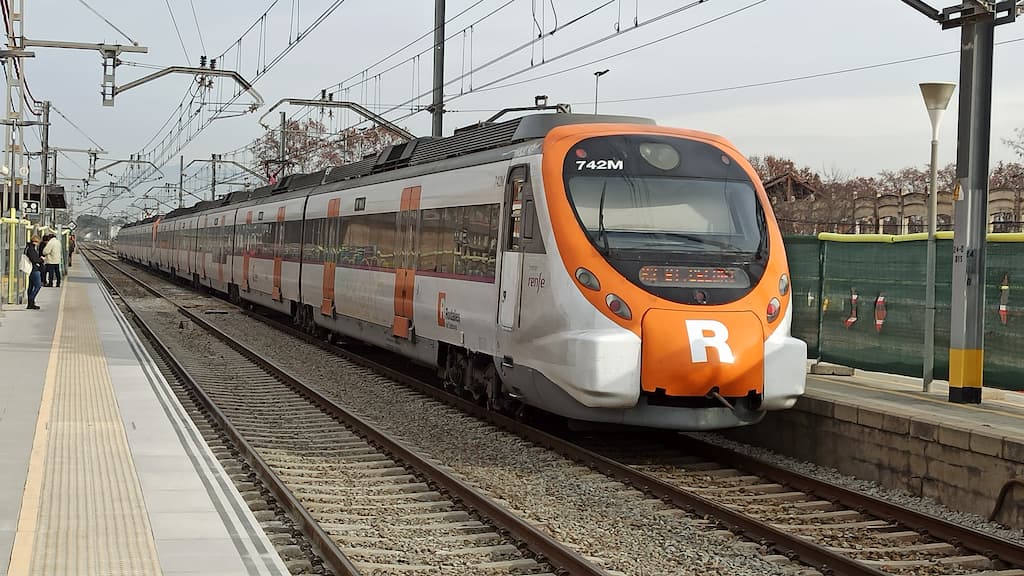 A white and orange Rodalies commuter train, labeled with the number 742M and the letter 'R', is stationed on the tracks at a platform. The train is part of the R2 North line, connecting Barcelona Airport to Sant Celoni. The platform is equipped with overhead electric lines, and a few passengers are visible in the background.
