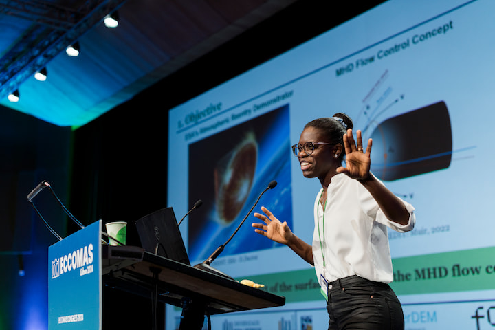 A speaker presents on stage at the ECCOMAS Congress 2024, organized by the CIMNE Congress Bureau. The speaker, wearing glasses and a white blouse, gestures with their hands while addressing the audience. Behind them, a large screen displays a slide titled 'MHD Flow Control Concept,' featuring a visual of fluid dynamics and a diagram of the concept. The podium bears the ECCOMAS Congress logo and the year 2024.