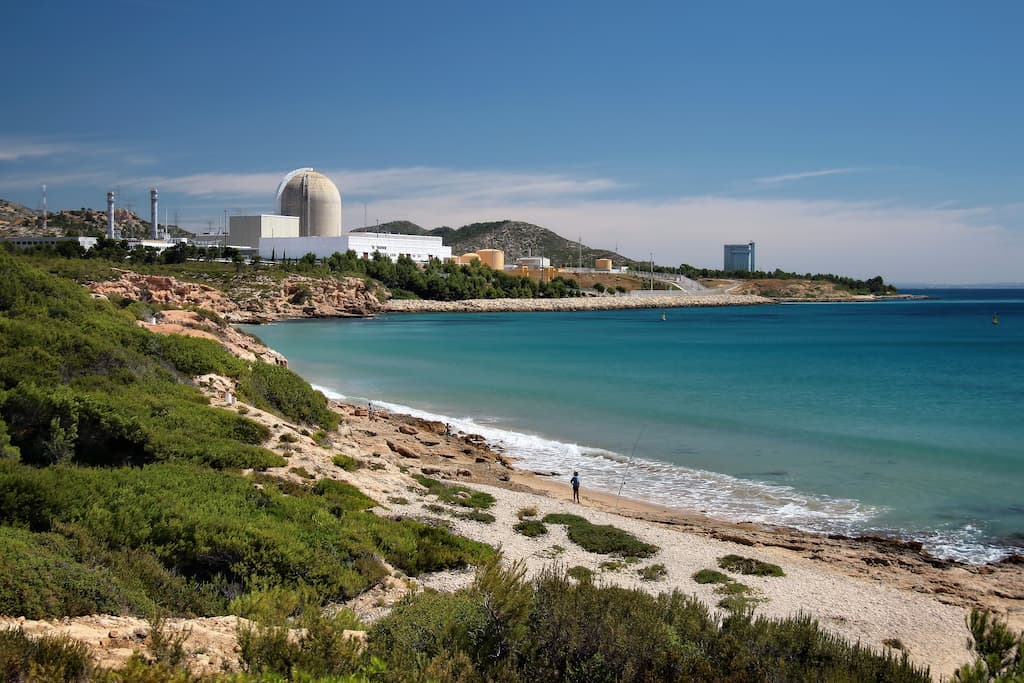 Panoramic view of the Vandellòs II Nuclear Power Plant, located on the Mediterranean coast near Vandellòs i l’Hospitalet de l’Infant in Catalonia, Spain. The image prominently features the plant’s large, domed containment building, which houses the pressurized water reactor (PWR). Surrounding the main reactor structure are various auxiliary buildings and cooling systems, essential for the plant’s operation. The facility is set against a backdrop of lush, green hills and a clear blue sky, with a sandy beach and the calm, turquoise waters of the Mediterranean Sea in the foreground. The contrast between the industrial complex and the natural coastal landscape highlights the plant’s strategic location, which allows it to use seawater for cooling purposes.