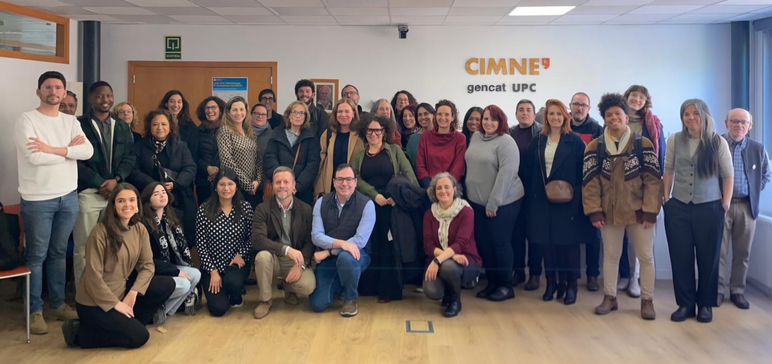 Group photograph of the first 2026 meeting of the CERCA centres Impact Community, held at the International Centre for Numerical Methods in Engineering (CIMNE) in Barcelona. The image shows a diverse group of people, including academic, research and research support staff, posing together in a room with the 'CIMNE gencat UPC' sign in the background. The participants are arranged in two rows: most are standing and some are seated or kneeling in the front row. Everyone is looking at the camera and some are smiling. The room has a professional atmosphere, with light-coloured walls and a door with a visible emergency exit sign.
