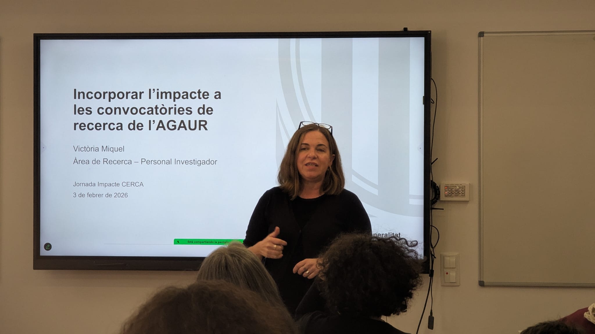 Photograph of Victòria Miquel, from the Research Area – Research Staff at AGAUR (Agency for Management of University and Research Grants), during her presentation entitled 'Incorporating impact into AGAUR research calls'. The image was taken on 3 February 2026 during the CERCA Impact Conference. Victòria Miquel is standing, speaking in front of a large screen showing the title of her presentation and her name. She is dressed in formal attire and glasses, and appears to be interacting with the audience. The setting is a conference room, with a whiteboard on the right.