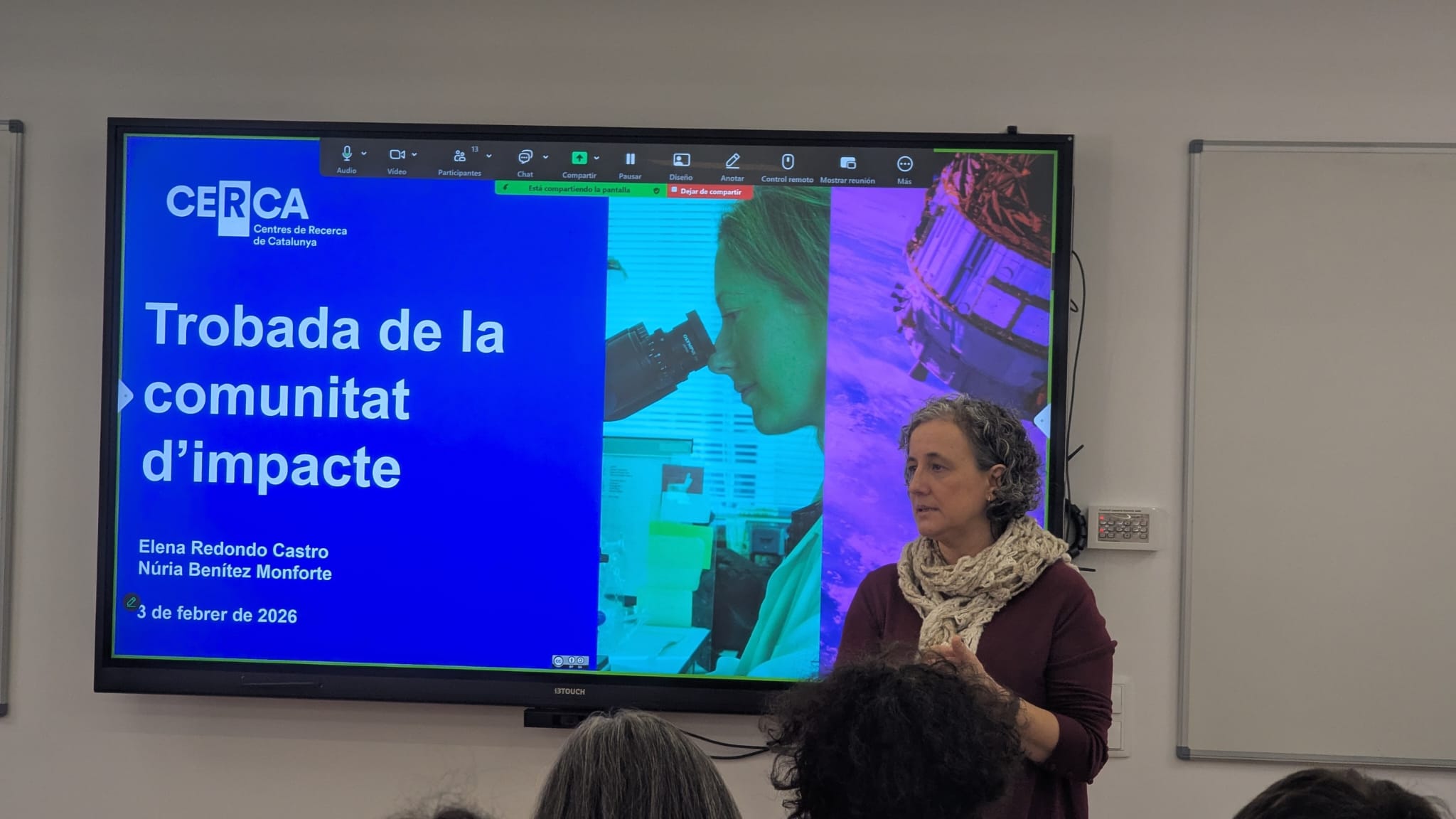 Photograph of Núria Benítez Monforte during her presentation at the 'Impact Community Meeting' of the CERCA Research Centres, held on 3 February 2026. Núria Benítez is standing, presenting in front of a large screen showing a slide with the title 'Impact Community Meeting' and her name alongside Elena Redondo Castro's. The slide includes a background image of a researcher using a microscope. Núria Benítez is wearing a long-sleeved T-shirt and a scarf, and appears to be interacting with the audience. The setting is a conference room, with a whiteboard on the right.