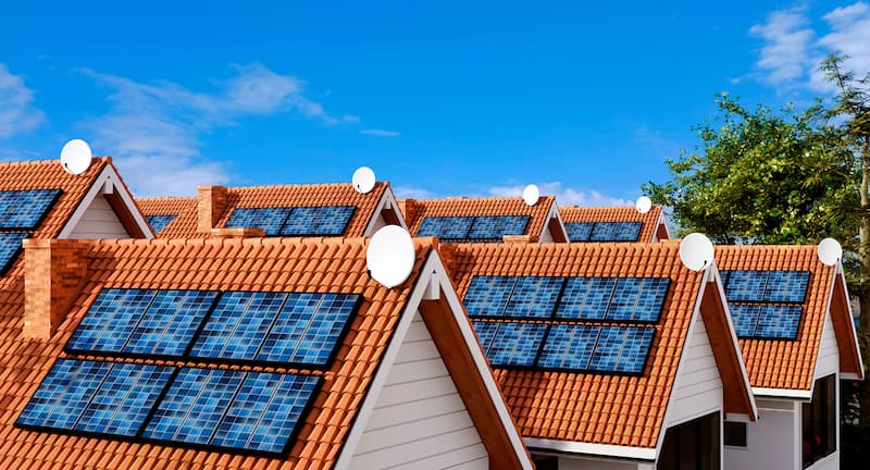 A row of modern houses with red-tiled roofs, each fitted with several solar panels on the sloped sections. White satellite dishes are mounted near the roof ridges. The sky is mostly clear with a few clouds, and a green tree stands on the right side of the scene.