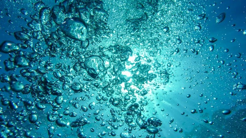 Underwater scene with sunlight filtering from above and numerous air bubbles rising toward the surface, creating a sense of depth and motion.