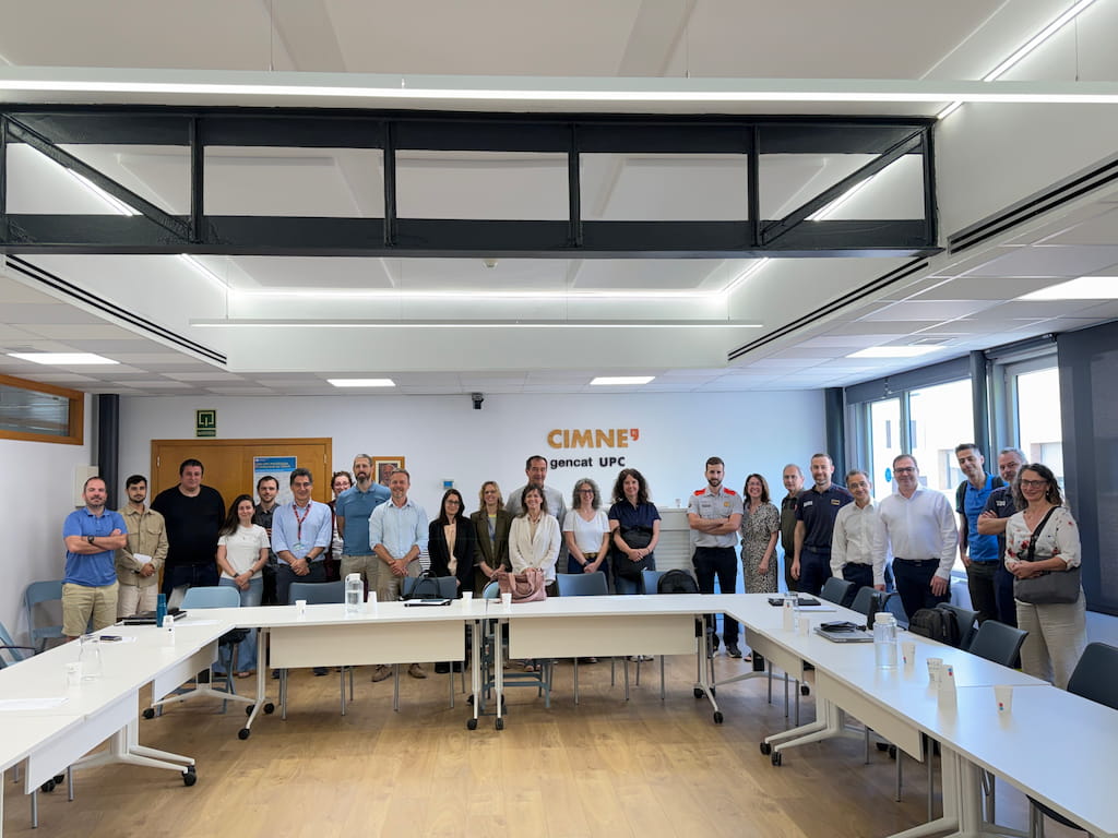 A group of around twenty people standing together in a bright meeting room at CIMNE’s headquarters in Barcelona. They are positioned behind a U-shaped arrangement of white tables, smiling and facing the camera. The wall behind them displays the CIMNE logo along with “gencat UPC,” indicating the collaboration between the International Centre for Numerical Methods in Engineering and the Catalan Government. The attendees include representatives from CIMNE and the Catalan Ministry of Home Affairs and Public Safety, some wearing uniforms from civil protection and emergency services. The modern room, with wooden floors and large windows, conveys a professional and collegial atmosphere following a productive working session on emergency management and civil protection.