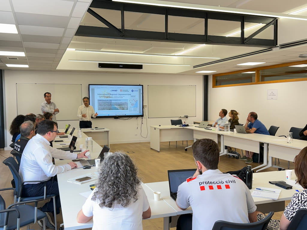 A meeting room at CIMNE’s headquarters in Barcelona where senior representatives from the International Centre for Numerical Methods in Engineering (CIMNE) and the Catalan Ministry of Home Affairs and Public Safety are gathered around a U-shaped table. Two presenters stand at the front near a large screen displaying a slide titled “Interacció amb el Seguretat i l’alertament,” featuring CIMNE and Protecció Civil logos, meteorological data, and a risk map. Attendees, including members of civil protection units—one wearing a shirt labeled “protecció civil”—listen attentively, taking notes and working on laptops. The bright, modern room reflects a collaborative atmosphere focused on emergency management and civil protection strategies.