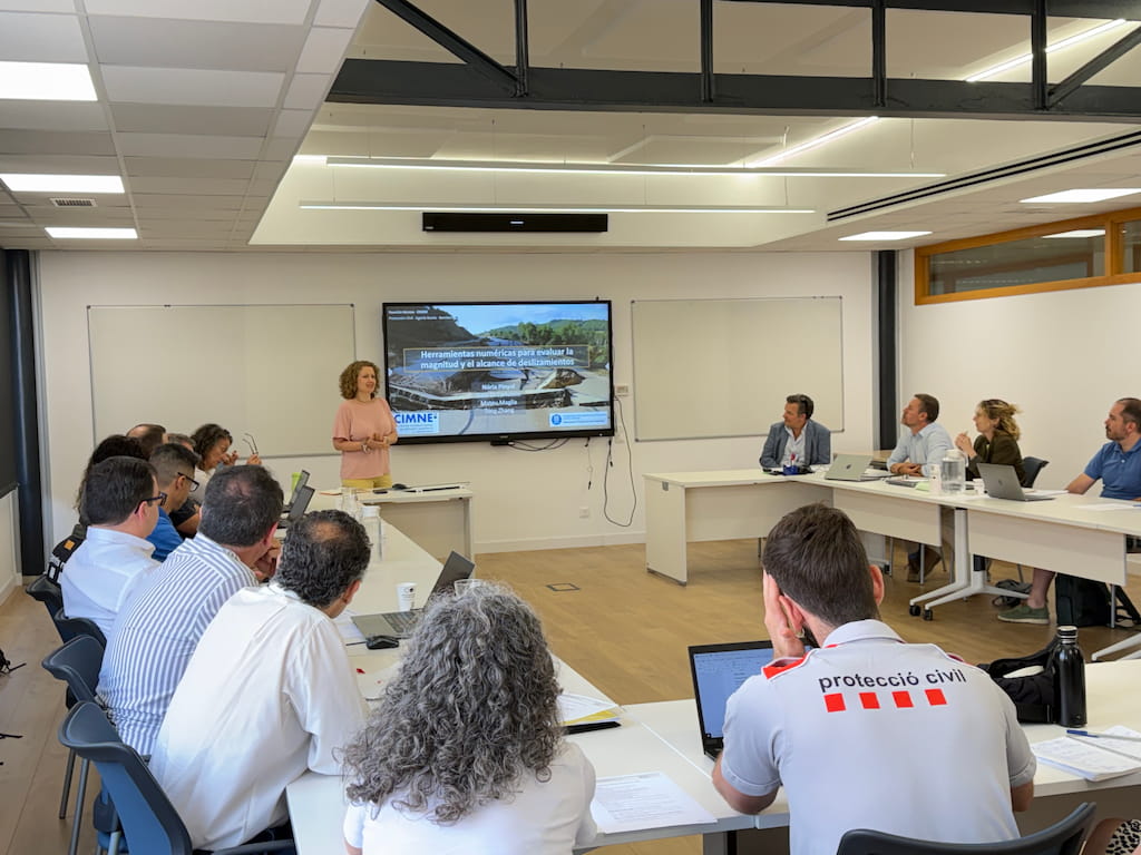 A professional meeting at CIMNE’s headquarters in Barcelona, showing a group of participants seated around a U-shaped table while a woman stands at the front giving a presentation. The slide on the large screen reads “Herramientas numéricas para evaluar la magnitud y el alcance de deslizamientos” and includes the names Nadia Pinyol and Mónica Mijares, along with the CIMNE logo and an image of a landslide area. Attendees from CIMNE and the Catalan Ministry of Home Affairs and Public Safety, including a participant wearing a “protecció civil” shirt, listen attentively and take notes on laptops. The bright, modern room conveys a collaborative atmosphere focused on civil protection and numerical modelling for disaster risk assessment.