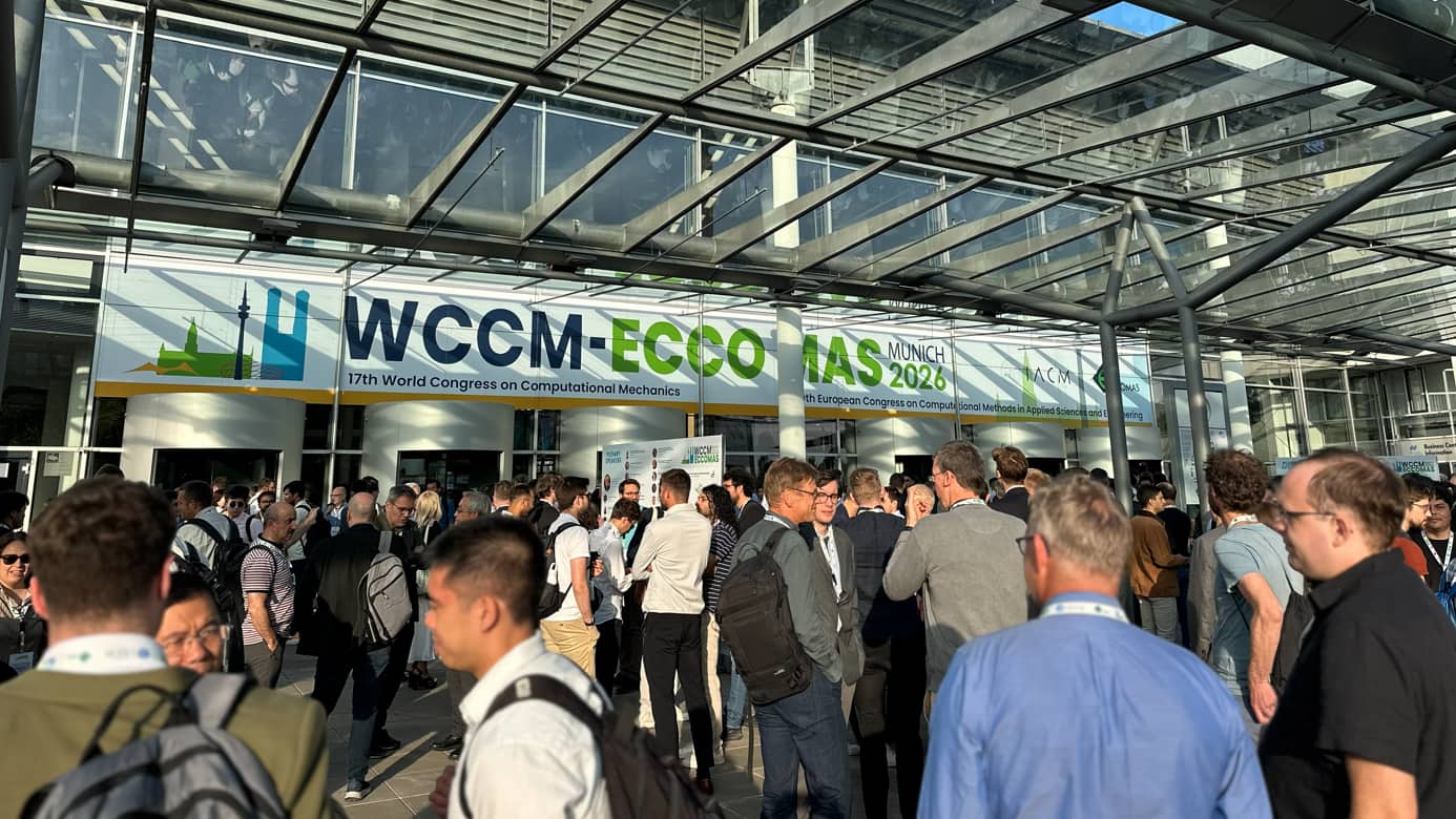  large group of conference attendees gathers outside a modern glass building under a transparent canopy, with a banner above the entrance announcing “WCCM·ECCOMAS Munich 2026,” the World Congress on Computational Mechanics and the European Congress on Computational Methods in Applied Sciences and Engineering.