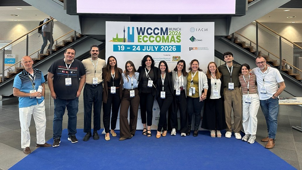 Twelve members of the conference secretariat team stand indoors in front of a large banner for WCCM·ECCOMAS Munich 2026, wearing conference badges. The banner displays the event dates, organizing societies, and Munich-themed graphics, creating a formal setting that highlights the team’s role in supporting the congress.