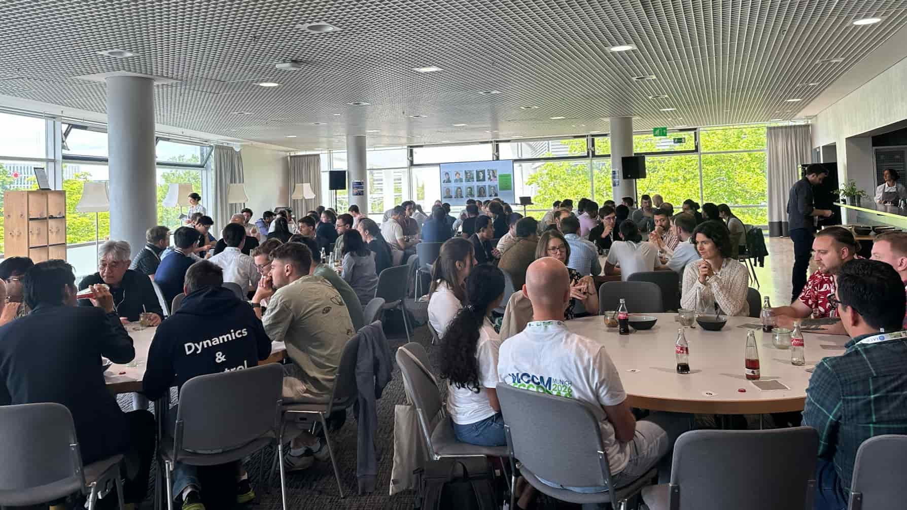 Participants sit at round tables during a conference meal or networking session in a bright, modern venue with large windows. Several attendees wear lanyards and shirts with event branding, and the room features posters and screens in the background.