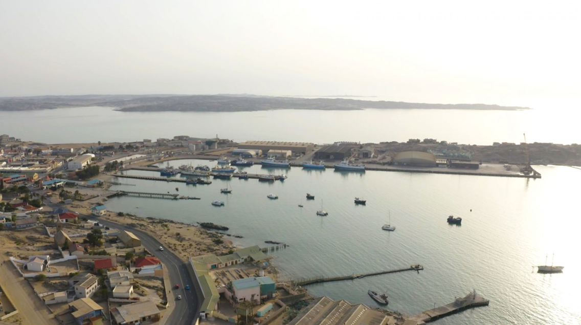 Vista aèria del port de Walvis Bay a Namíbia, que mostra aigües tranquil·les salpicades d'embarcacions de pesca i petits vaixells a prop dels molls. La zona portuària inclou magatzems, edificis industrials i carreteres costaneres que connecten amb el paisatge arenós de la ciutat. A la llunyania, la llacuna i la península s'estenen sota un cel boirós, il·lustrant l'entorn costaner àrid de la regió i la seva infraestructura marítima.  