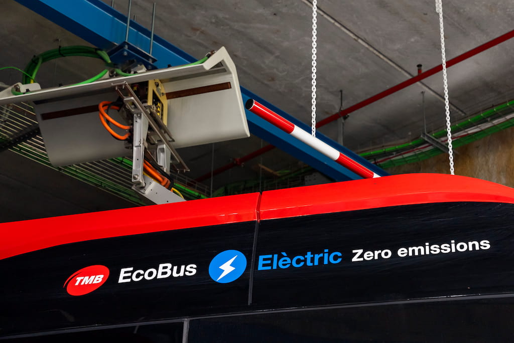 Primer pla d’un autobús elèctric de TMB en un espai de manteniment, amb el pantògraf connectat al sistema de recàrrega superior. A la carrosseria negra i vermella s’hi llegeix «TMB EcoBus – Elèctric – Zero emissions». La imatge il·lustra l’electrificació de la flota d’autobusos de Barcelona i la transició cap a un transport públic amb menys emissions. Foto: Ajuntament de Barcelona sota llicència CC BY-NC-ND 2.0